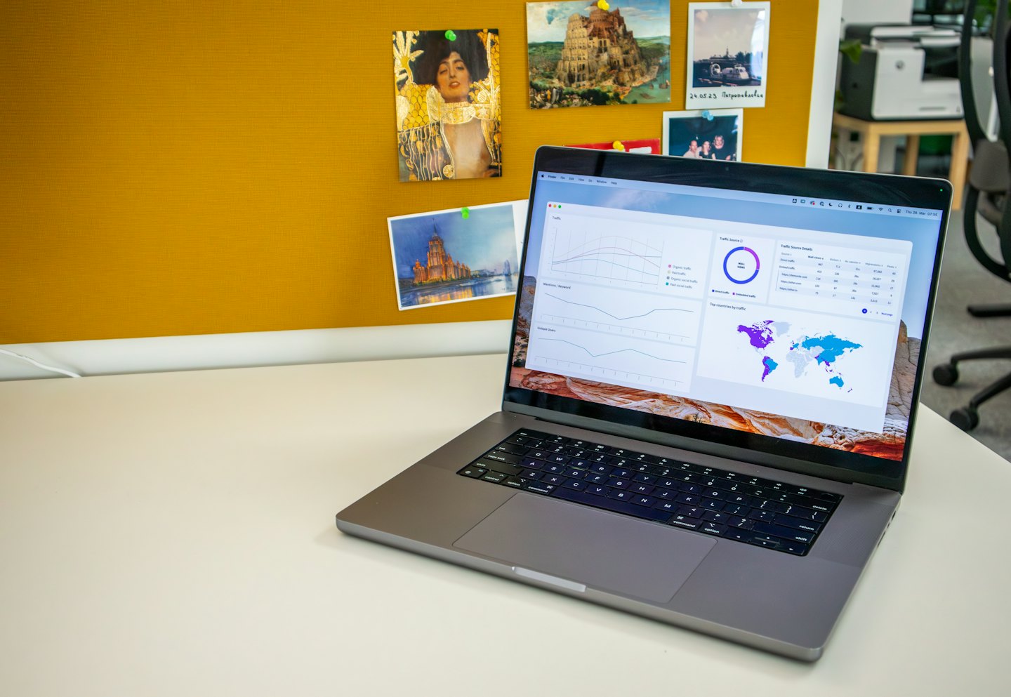 Laptop displaying charts on a white desk with a yellow corkboard.